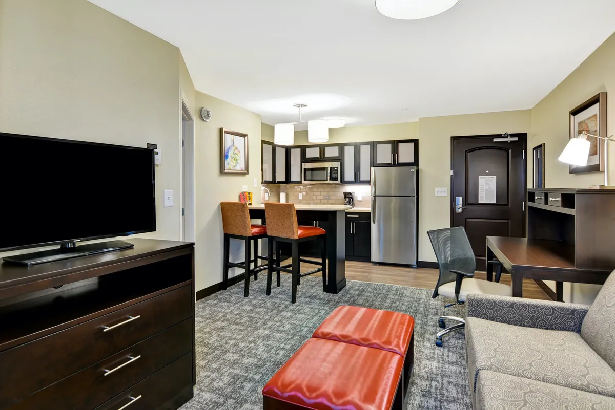 The image showcases a welcoming, open-concept living space with a fully equipped kitchen, cozy dining area, plush sofa facing a TV, a sleek ottoman, and a well-lit desk by the window—inviting guests to cook, dine, relax, and work comfortably.