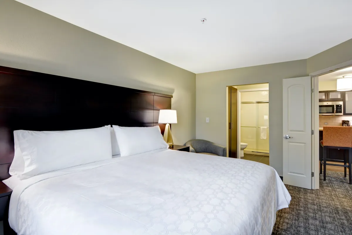 This image captures a cozy interior featuring a king-size bed, a lamp, the entrance to a bathroom, and, importantly, a kitchenette area. The small table set up near the kitchen hints at enjoying meals or snacks in the comfort of your room. Appliances and kitchen essentials are visible, providing a glimpse of the full kitchen setup available to guests.