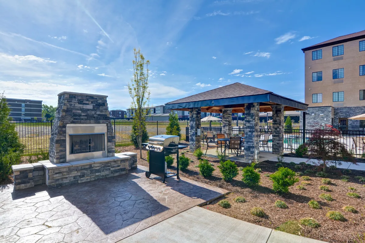 An inviting exterior patio featuring a stone fireplace, a modern grill, a gazebo surrounded by lush landscaped greenery, and comfortable seating for outdoor gatherings. The space exudes a friendly, welcoming atmosphere ideal for socializing or winding down after a productive day.