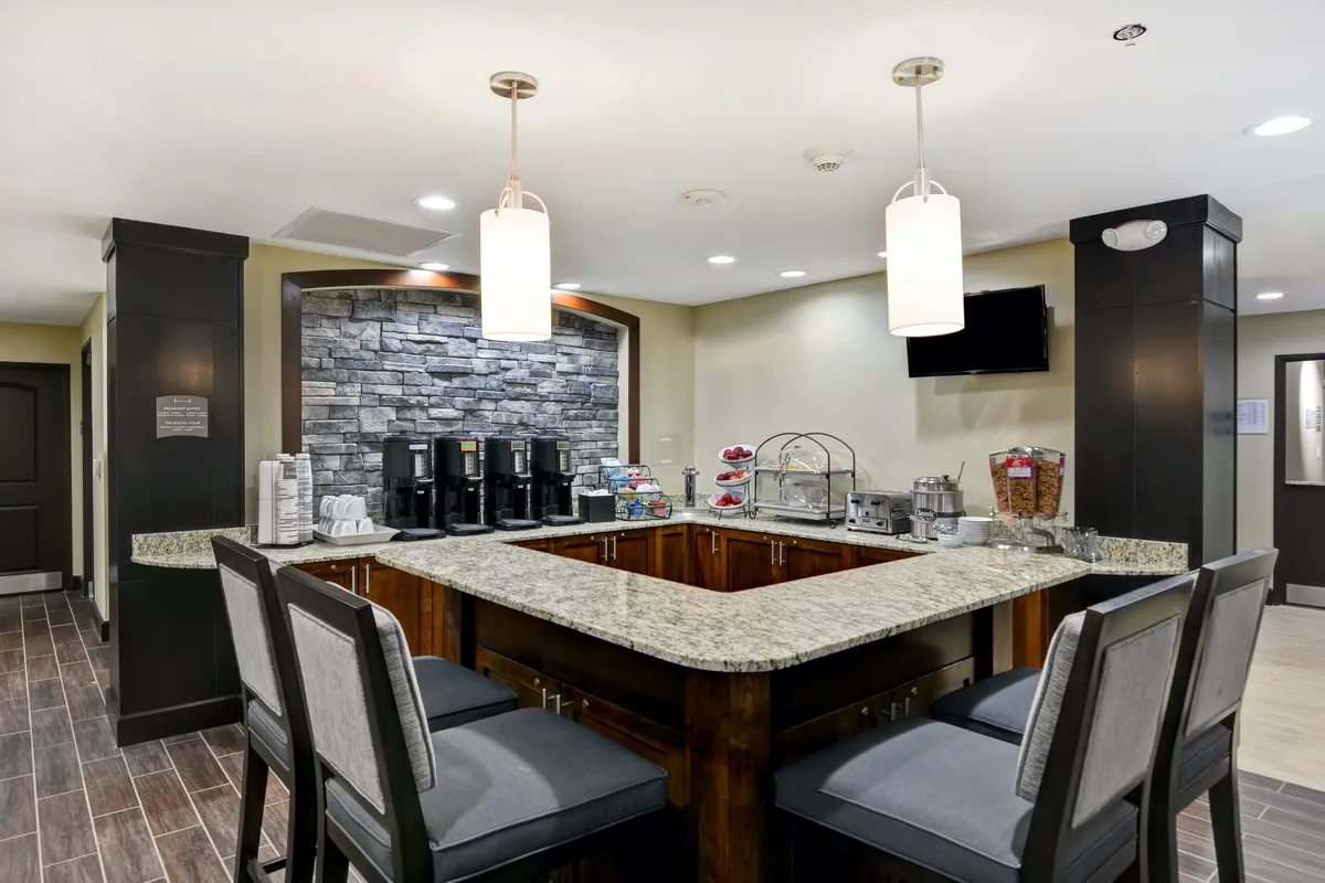 A welcoming interior scene featuring a granite countertop coffee and beverage station, complemented by a stone accent wall and casual seating areas. Guests are seen enjoying coffee, planning their day, and exchanging friendly local recommendations.