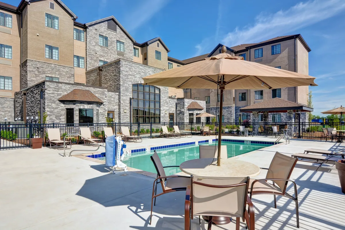 A vibrant outdoor swimming pool area with inviting lounge chairs, umbrellas, and the hotel building in the background. Guests are seen swimming, sunbathing, and socializing, all set against a warm sunset backdrop.