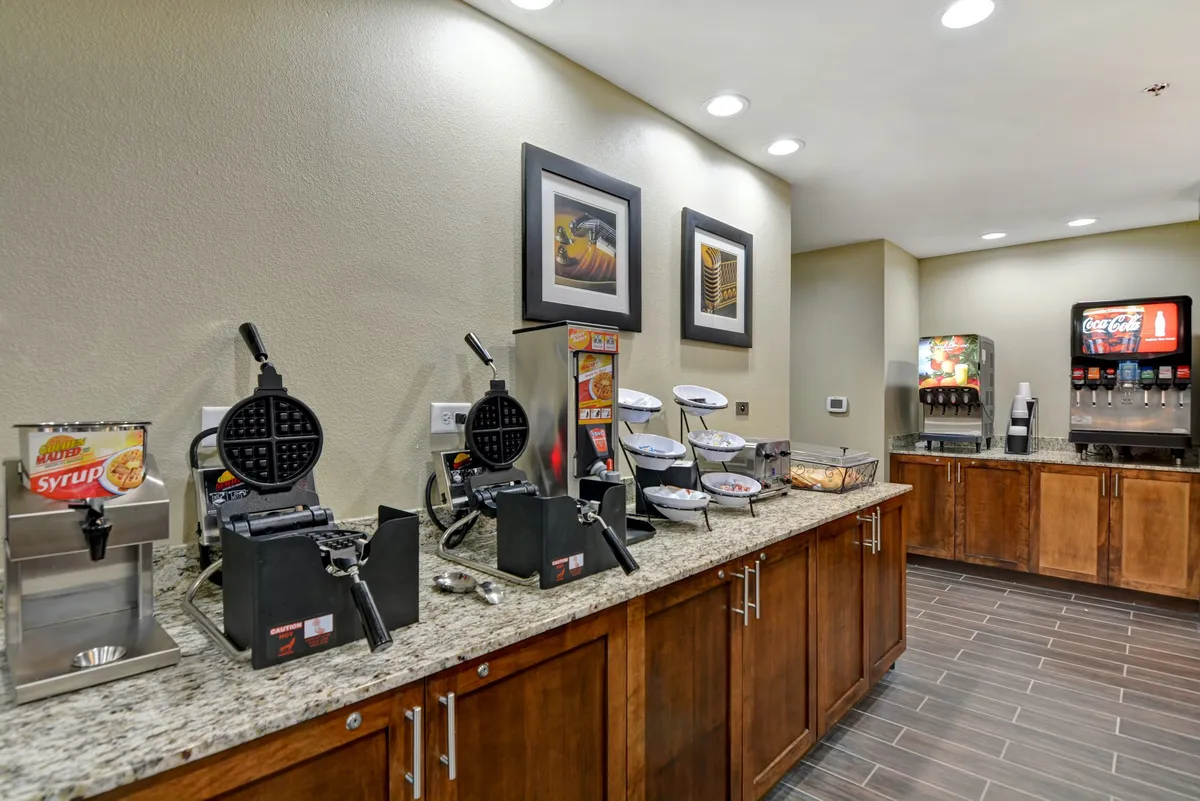 This image features a waffle maker, syrup dispenser, and a beverage station, with decorative wall art in the background. The setup indicates a communal or complimentary breakfast area where guests can quickly prepare breakfast or enjoy fresh options each morning.