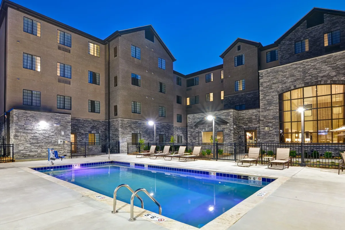 An inviting outdoor swimming pool with clear blue water sits at the center, surrounded by comfortable lounge chairs and the condo's building facade in the background. The scene conveys a relaxed, social atmosphere, ideal for enjoying summer days outdoors.