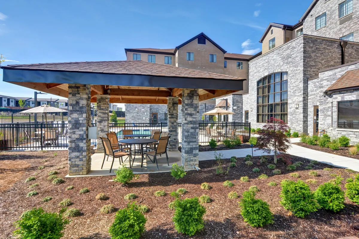 A sunny outdoor dining area under a stone gazebo adjacent to a landscaped garden, with a swimming pool in the background. The scene is set for casual meals, social gatherings, or a peaceful moment outdoors.